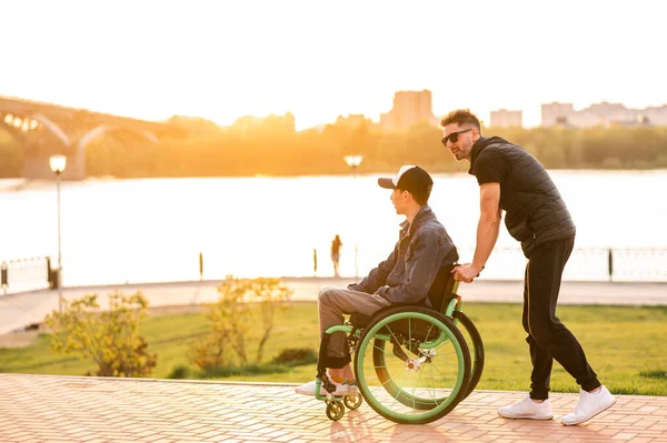 Человек в инвалидном кресле гуляет со своим другом. Человек, гуляющий с другом-инвалидом в инвалидном кресле в парке — стоковое фото
