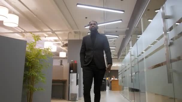 Retrato de joven empresario afroamericano guapo y confiado en traje formal caminando por el pasillo de la oficina moderna para reunirse con el cliente usando el ordenador portátil — Vídeos de Stock