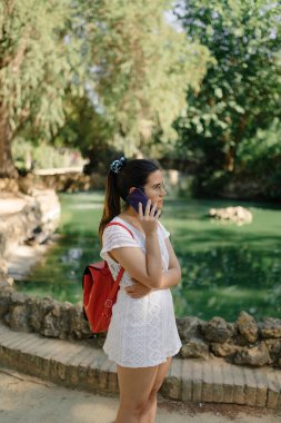 Beyaz elbiseli, kırmızı sırt çantalı, parkta telefonla konuşan genç bir kadının orta boy fotoğrafı.