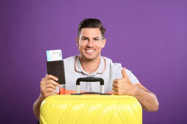 Pasaportunda mor arka planda yaz gezisi için bavulu ve bileti olan yakışıklı bir adam. Tatil gezisi