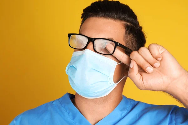 黄色の背景に医療用マスクを身に着けていることによって引き起こされる霧の眼鏡を拭く医師 クローズアップ — ストック写真
