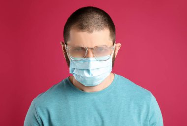 Pembe arka planda tek kullanımlık maske takan sisli gözlüklü bir adam. Koronavirüs salgını sırasında koruyucu tedbir