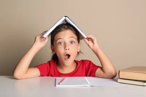 Emocjonalna Dziewczynka Książką Głowie Odrabiająca Lekcje Przy Stole Beżowym Tle — Zdjęcie stockowe
