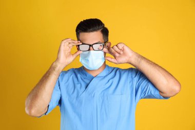 Doktor sisli camları siliyor. Sarı arka planda tıbbi maske takıyor.