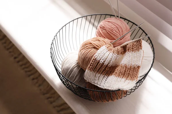 Yarn balls and knitting needles in metal basket on window sill indoors. Creative hobby