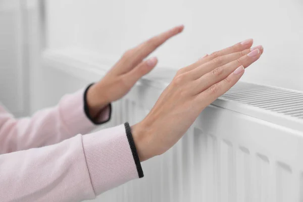 Woman warming hands on heating radiator near white wall, closeup