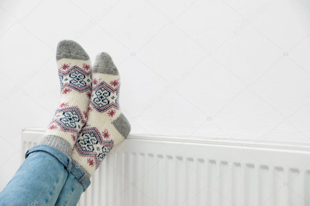 Woman warming legs on heating radiator near white wall, closeup. Space for text