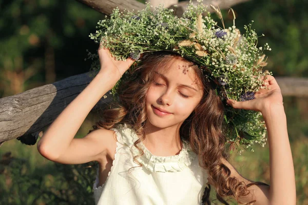 晴れた日に屋外の木製のフェンスの近くに美しい花で作られた花輪を着てかわいい女の子 — ストック写真