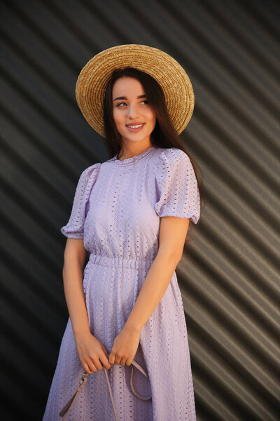 Beautiful young woman in stylish violet dress and straw hat near dark corrugated metal wall