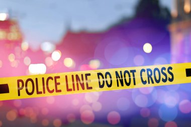 Sarı emniyet şeridi olay yerini izole ediyor. Şehrin bulanık görüntüsü, kırmızı ve mavi polis arabası ışıkları.