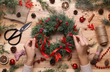 Çiçekçi güzel bir Noel çelengi yapıyor, böğürtlenli ve kırmızı kurdeleli, ahşap masa manzaralı.