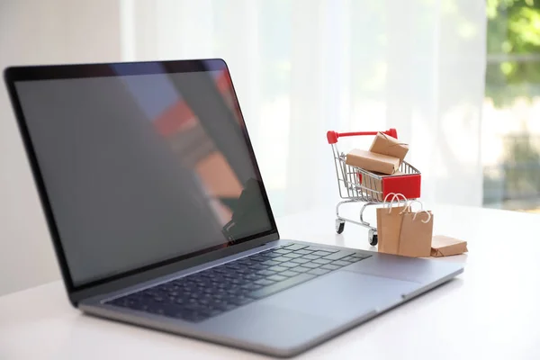 Internet shopping. Modern laptop, small cart with boxes and bags on table indoors