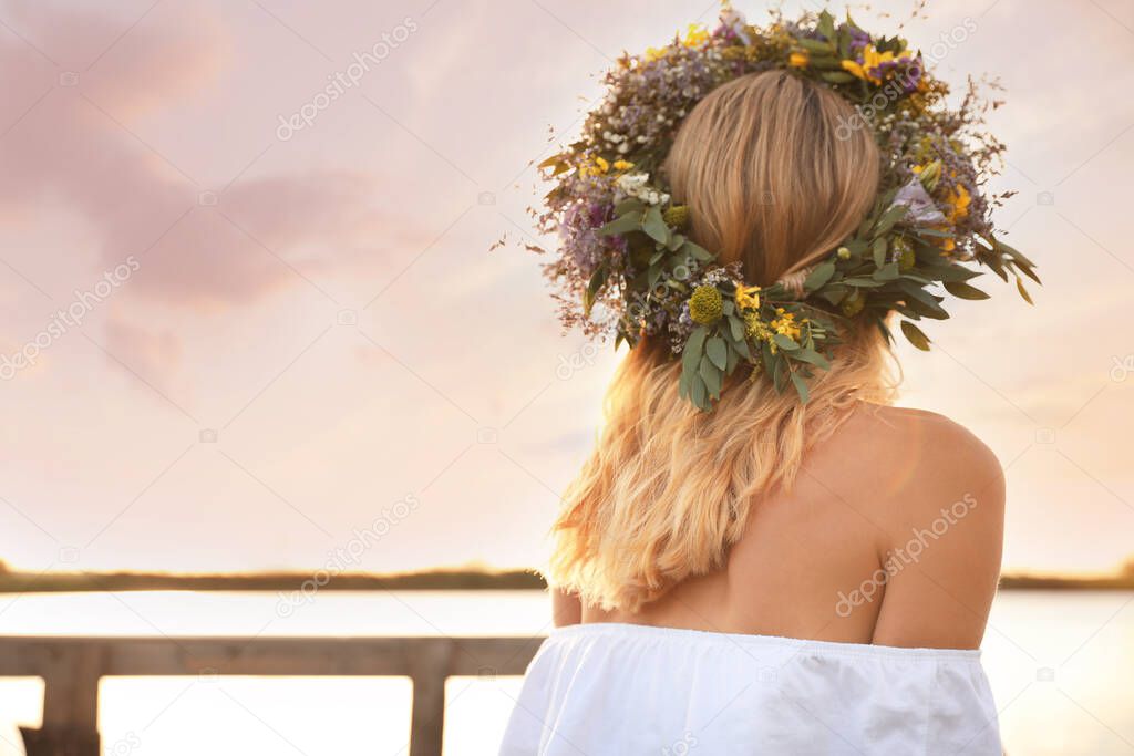 Young woman wearing wreath made of beautiful flowers near river on sunny day