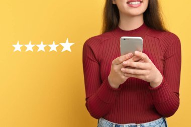 Sarı arka plandaki akıllı telefondan beş yıldızlı gözden geçirme sitesinden ayrılan bir kadın, yakın plan. Servis veya ürün geri bildirimi