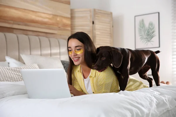 Young woman with eye patches working on laptop near her dog in bedroom. Home office concept