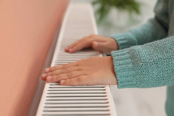 Child warming hands on heating radiator indoors, closeup