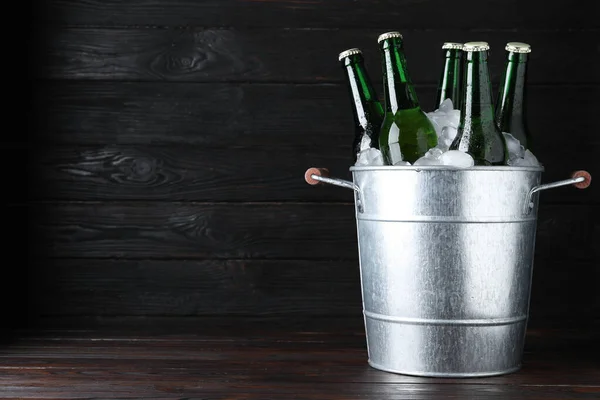 Metal bucket with bottles of beer and ice cubes on wooden table against dark background, space for text