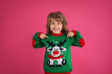 Şirin küçük kız pembe arka planda yeşil Noel süveterini gösteriyor.