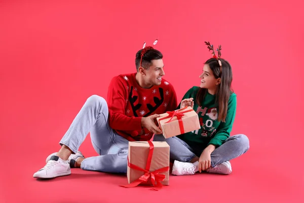 漂亮而快乐的夫妇 戴着圣诞头巾和毛衣 戴着红色背景的礼物坐着 — 图库照片