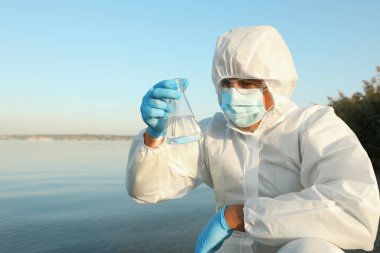 Kimyasal koruyucu elbise giyen bilim adamı konik matarayla analiz için nehirden örnek alıyor.