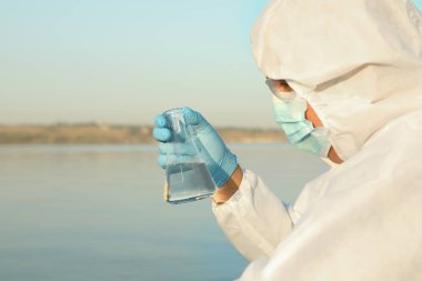 Kimyasal koruyucu elbise giyen bilim adamı konik matarayla analiz için nehirden örnek alıyor.