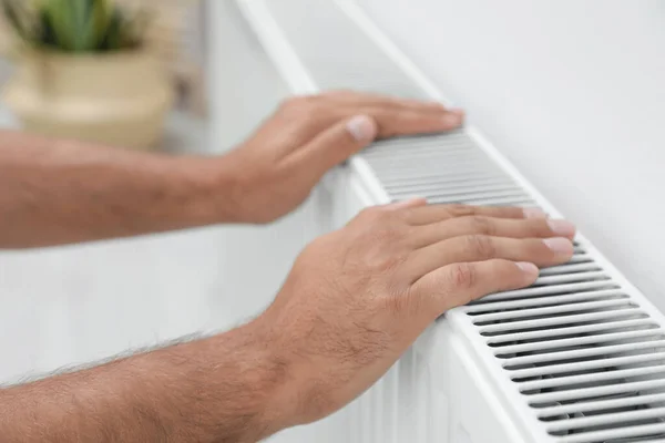 Man warming hands on heating radiator near white wall, closeup