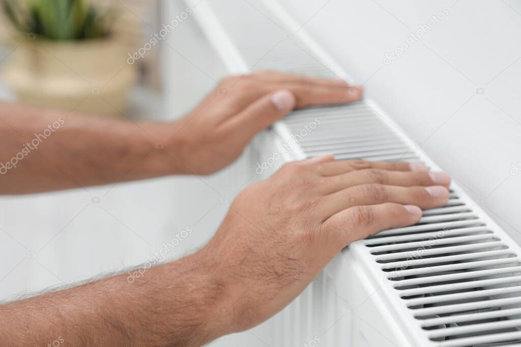 Man warming hands on heating radiator near white wall, closeup