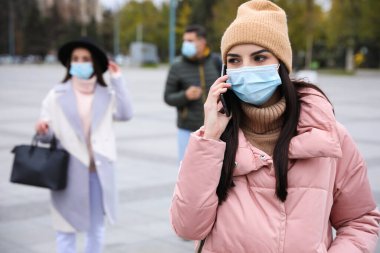 Sağlık maskeli genç bir kadın dışarıda yürürken telefonla konuşuyor. COVID-19 salgını sırasında kişisel koruma