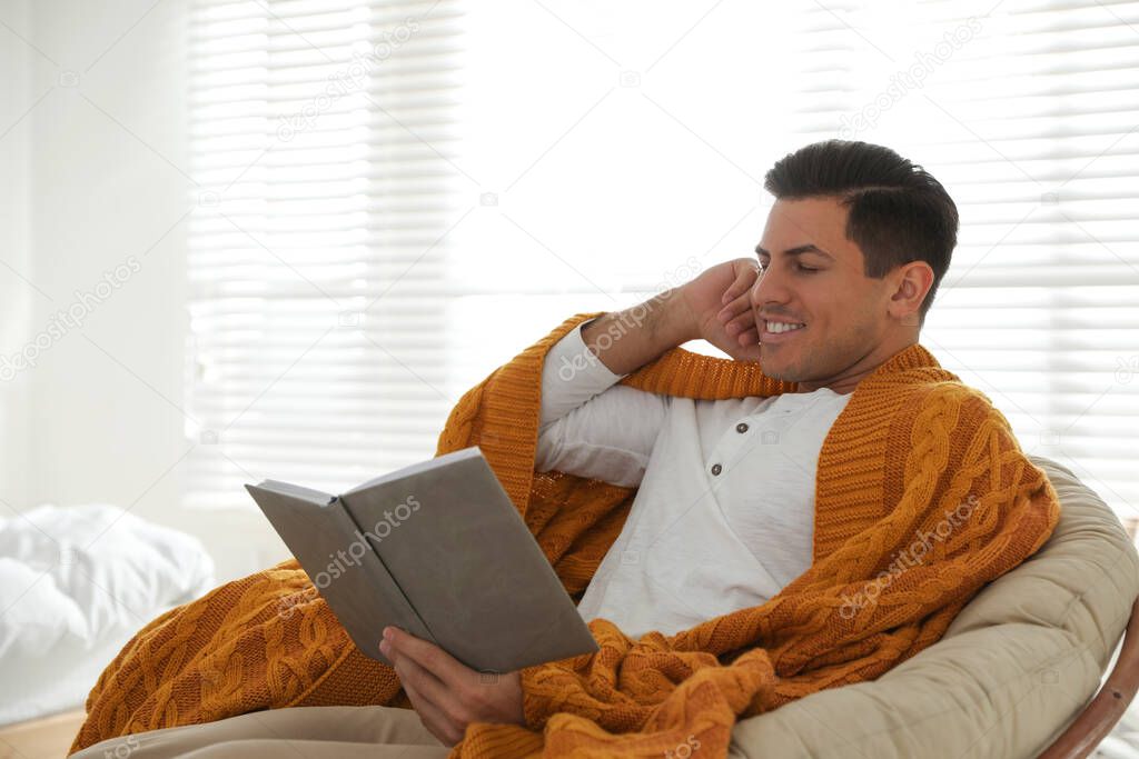 Man covered with warm orange plaid reading book at home