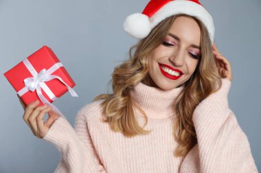 Mutlu genç bir kadın Noel Baba şapkası takıyor ve gri arka planda Noel hediyesi.