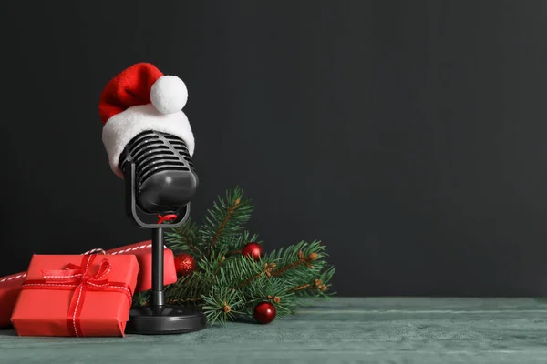 Retro microphone with Santa hat, gift boxes and festive decor on wooden table against black background, space for text. Christmas music