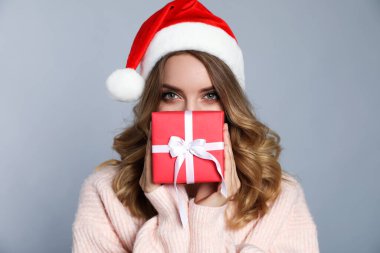 Mutlu genç bir kadın Noel Baba şapkası takıyor ve gri arka planda Noel hediyesi.