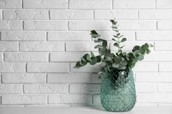 Beautiful eucalyptus branches in glass vase on white wooden table near brick wall. Space for text