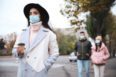 Dışarıda yürüyen bir fincan kahveyle tıbbi maske takan genç bir kadın. COVID-19 salgını sırasında kişisel koruma