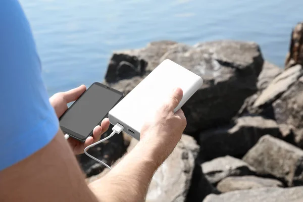 Man charging mobile phone with power bank on rocky mountain near river, closeup