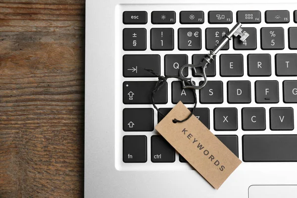 Laptop, vintage key and tag with word KEYWORDS on wooden table, top view