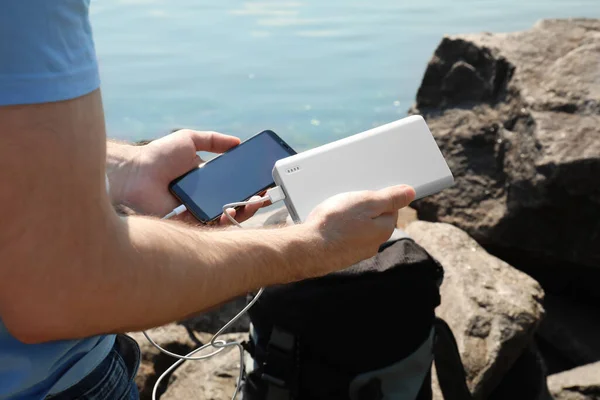 Man charging mobile phone with power bank on rocky mountain near river, closeup