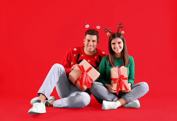 美しい幸せなカップルでクリスマスのヘッドバンドとセーター座ってギフトで赤の背景 — ストック写真
