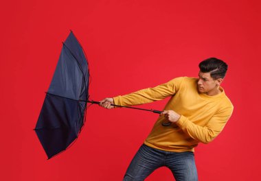 Emotional man with umbrella caught in gust of wind on red background clipart