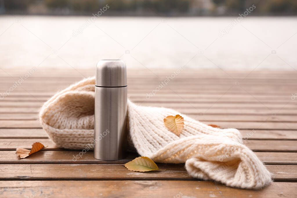 Metallic thermos, knitted scarf and fallen leaves on wooden pier near river. Space for text
