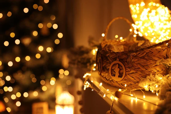 Fireplace mantel with wicker basket, festive lights and blurred Christmas tree on background