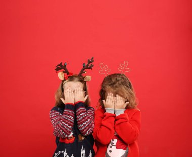 Kids in Christmas sweaters and festive headbands hiding faces on red background