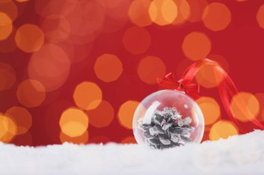 Beautiful transparent Christmas ornament with fir cone on snow against red background, bokeh effect. Space for text. Space for text