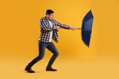 Emotional man with umbrella caught in gust of wind on yellow background