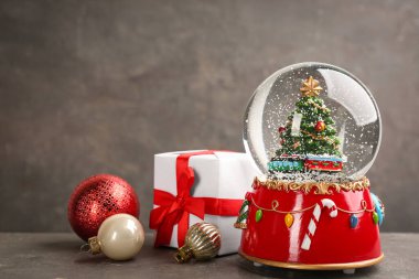 Güzel kar küresi, hediye kutusu ve gri masadaki Noel topları.