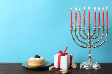 Silver menorah near dreidels with He, Pe, Nun, Gimel letters, gift box and sufganiyah on wooden table, space for text. Hanukkah symbols clipart