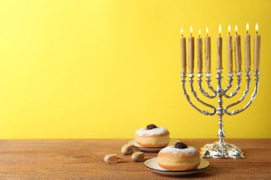 Gümüş menorah, dreidels with He, Pe, Nun, Gimel letters and sufganiyot on wood table against yellow background, space to text. Hanuka sembolleri