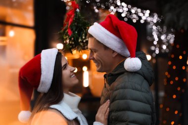 Noel Baba şapkalı mutlu çift dışarıda ökseotunun altında duruyor.