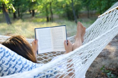 Yeşil bahçedeki hamakta kitap okuyan genç bir kadın.