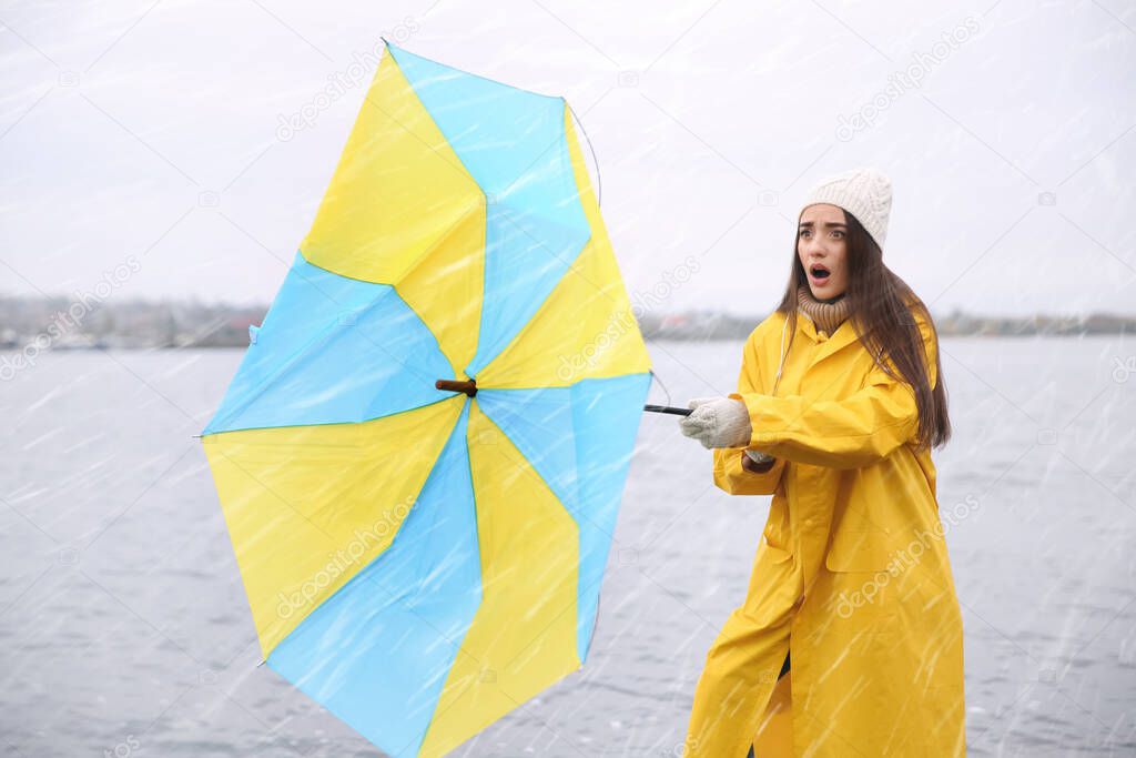 Woman in yellow raincoat with umbrella caught in gust of wind near river